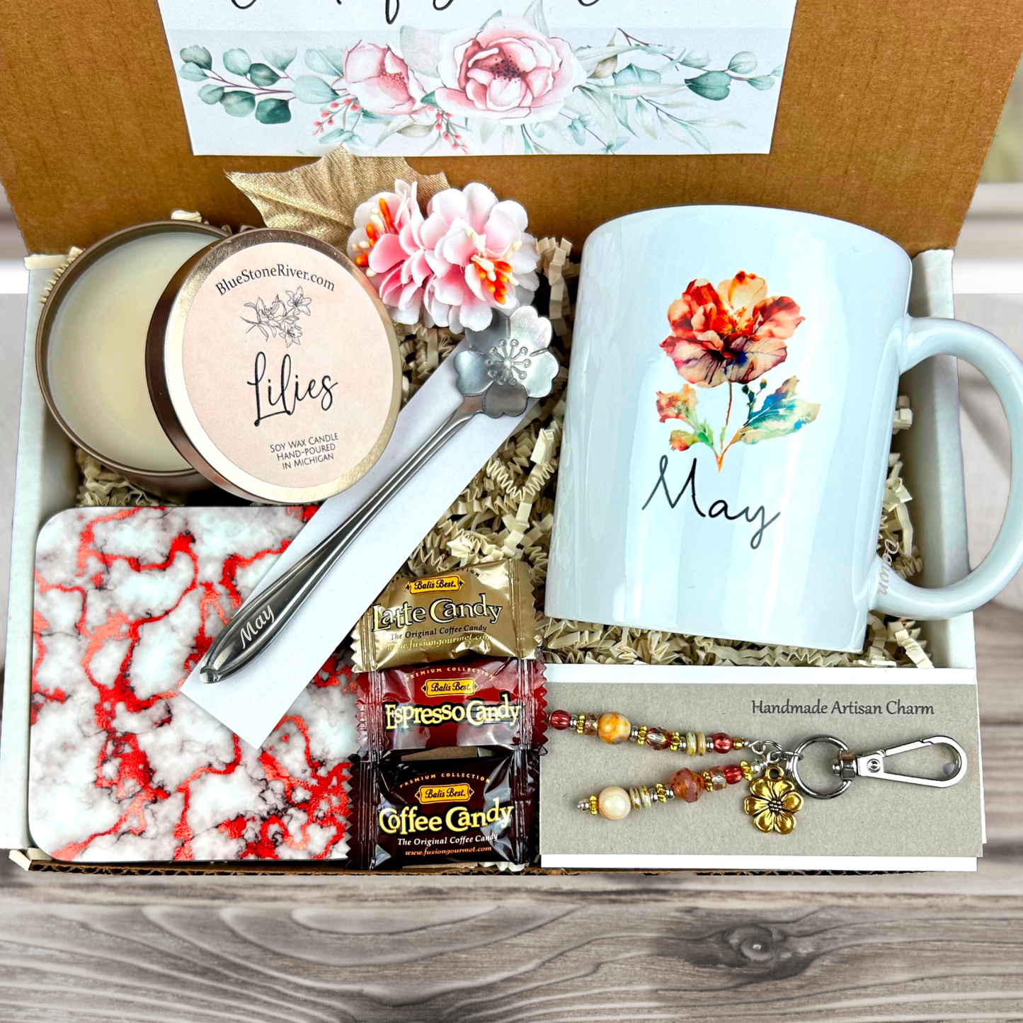 Unboxing view of complete gift basket with personalized floral mug, coaster, candy, artisan charm and scented candle.